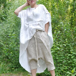 K&ouml;nnte beinhalten: Eine Frau tr&auml;gt ein wei&szlig;es Leinenoberteil mit R&uuml;schenkragen und eine beige Leinenhose mit Kordelzug. Sie tr&auml;gt eine wei&szlig;e Sonnenbrille und gelbe Sandalen.