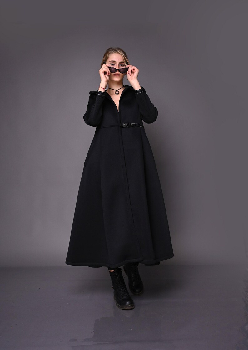 A model facing forward while adjusting black sunglasses, wearing a floor-length black coat and black boots.