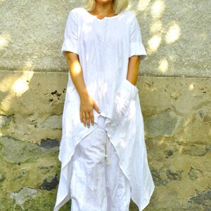 A woman in a white linen tunic and pants set, with a colorful fabric necklace, stands with her hands clasped in front of her.