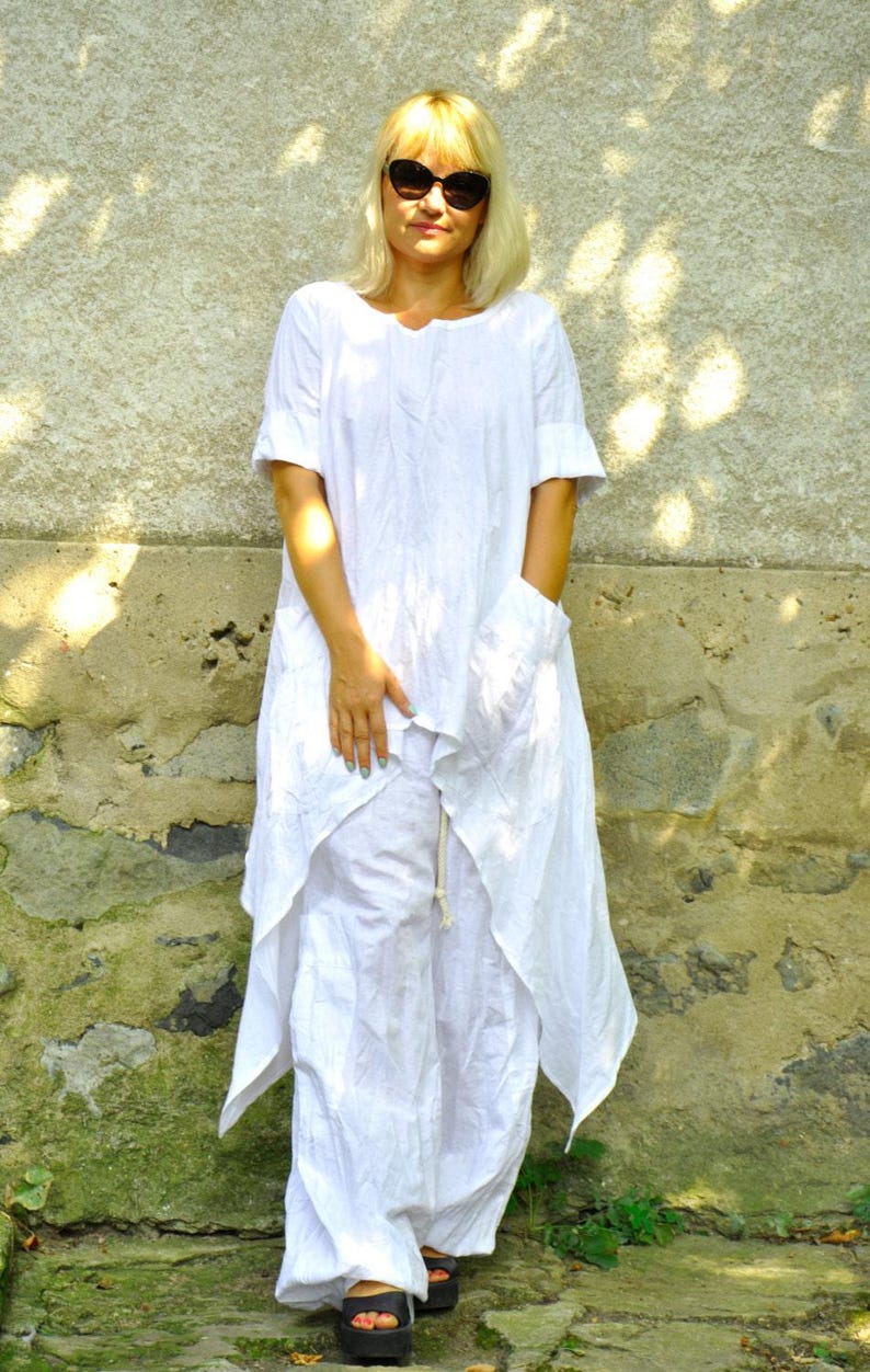 A woman in a white linen tunic and pants set, with a colorful fabric necklace, stands with her hands clasped in front of her.
