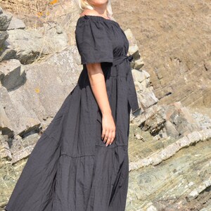 May include: A woman wearing a black maxi dress with a ruffled hem and a belt. She is standing on a rocky surface and wearing blue sunglasses and black sandals.
