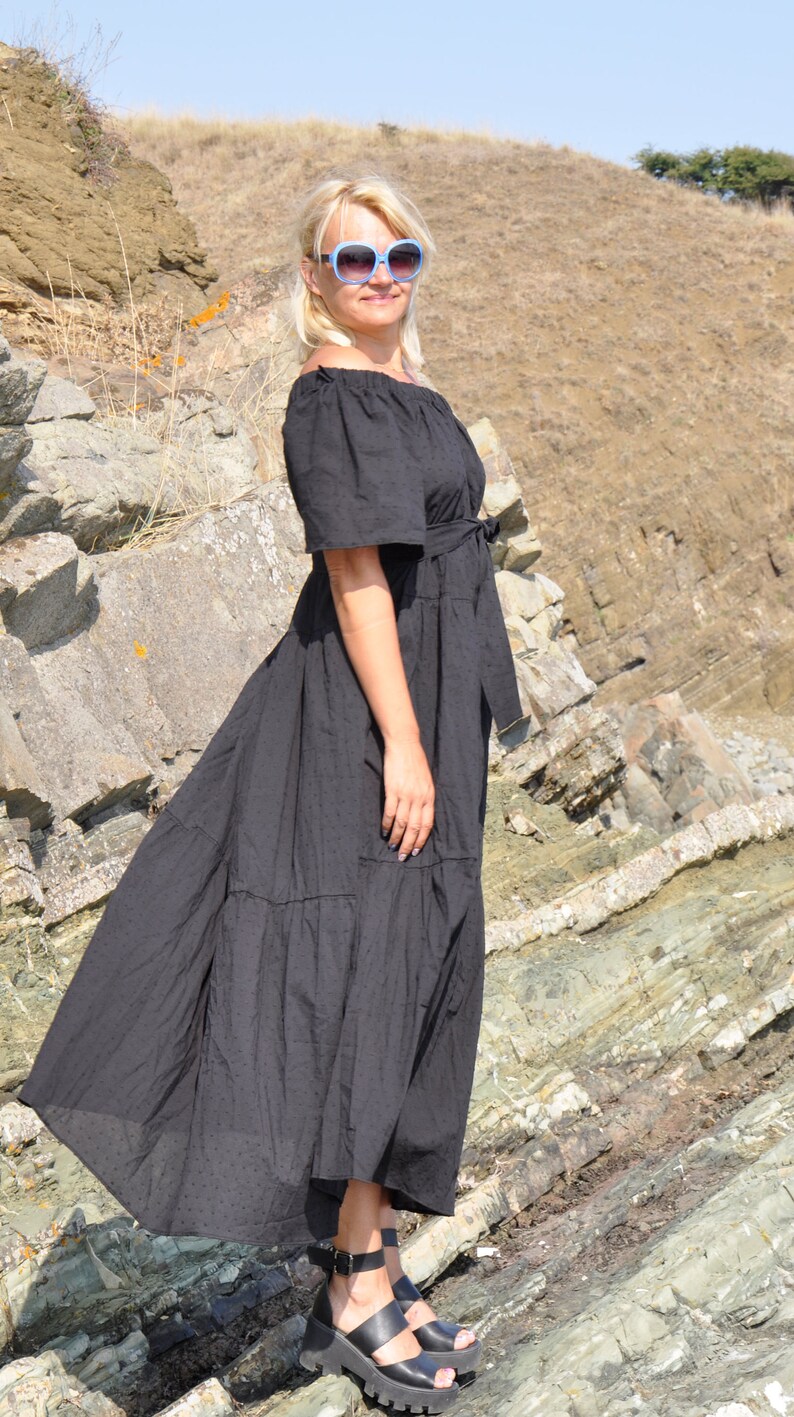 May include: A woman wearing a black maxi dress with a ruffled hem and a belt. She is standing on a rocky surface and wearing blue sunglasses and black sandals.