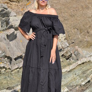 May include: A woman wearing a black off-the-shoulder maxi dress with a tiered skirt and a belt. She is wearing sunglasses and standing on a rocky surface.