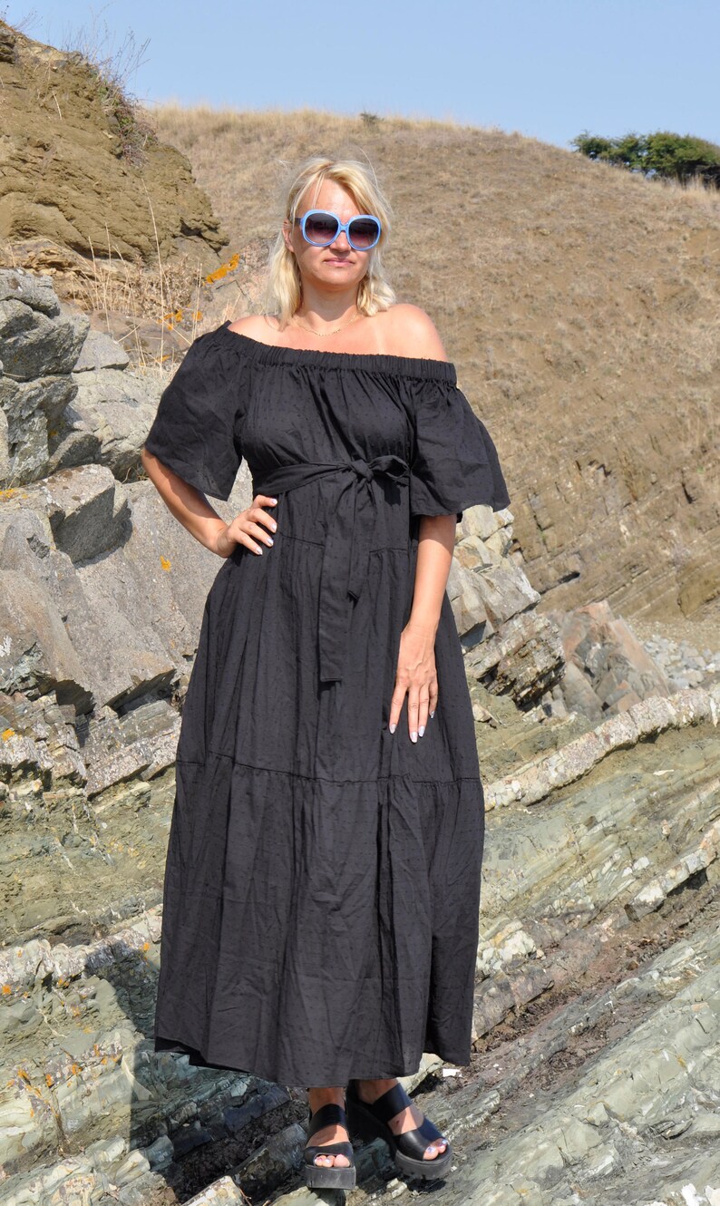May include: A woman wearing a black off-the-shoulder maxi dress with a tiered skirt and a belt. She is wearing sunglasses and standing on a rocky surface.