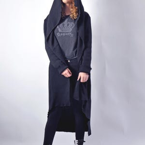 A full-body shot of a model facing forward, wearing a black hooded cardigan, grey graphic t-shirt, black pants, and white combat boots.