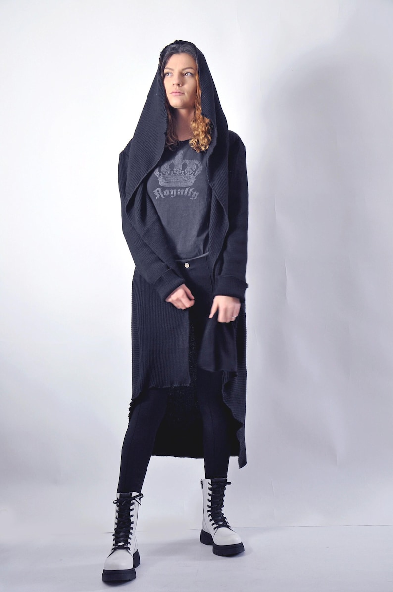 A full-body shot of a model facing forward, wearing a black hooded cardigan, grey graphic t-shirt, black pants, and white combat boots.