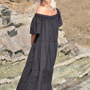 May include: A woman wearing a black off-the-shoulder maxi dress with a tiered skirt. She is standing on a rocky surface and looking away from the camera.
