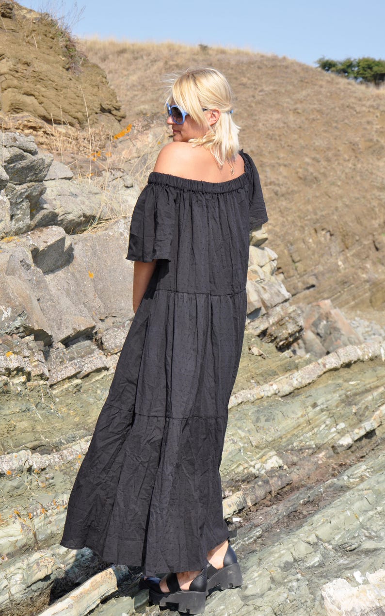 May include: A woman wearing a black off-the-shoulder maxi dress with a tiered skirt. She is standing on a rocky surface and looking away from the camera.