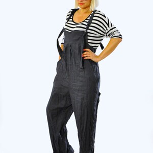 May include: Dark gray linen overalls with adjustable straps. The overalls have a relaxed fit and feature a pleated front. The model is wearing a black and white striped long-sleeve shirt and black platform sandals. The overall style is casual and comfortable.