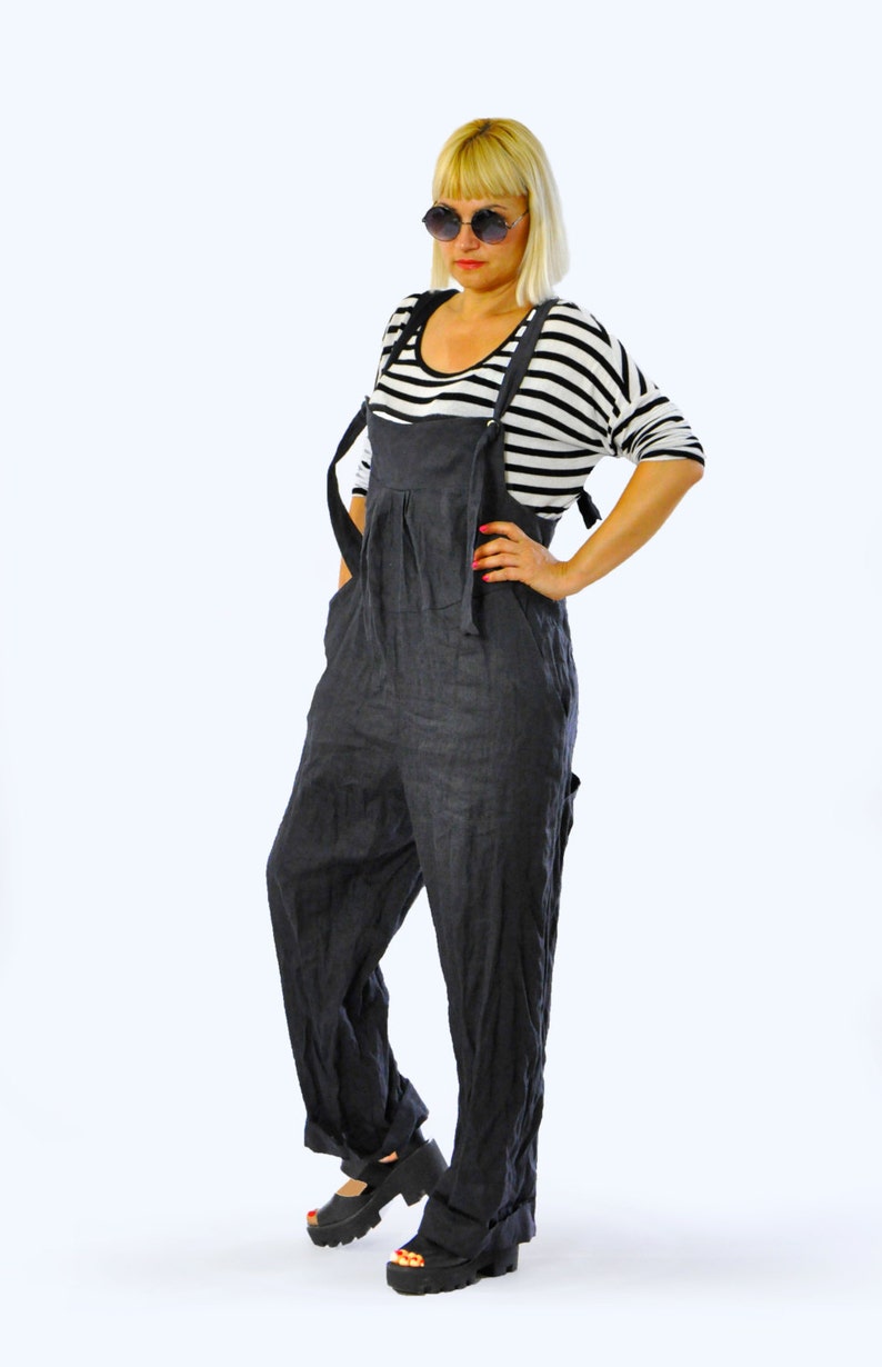 May include: Dark gray linen overalls with adjustable straps. The overalls have a relaxed fit and feature a pleated front. The model is wearing a black and white striped long-sleeve shirt and black platform sandals. The overall style is casual and comfortable.