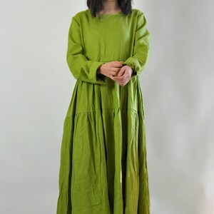 May include: A long, tiered, green linen dress with a round neckline and long sleeves. The dress is loose-fitting and has a flowy silhouette. The model is wearing white sneakers.
