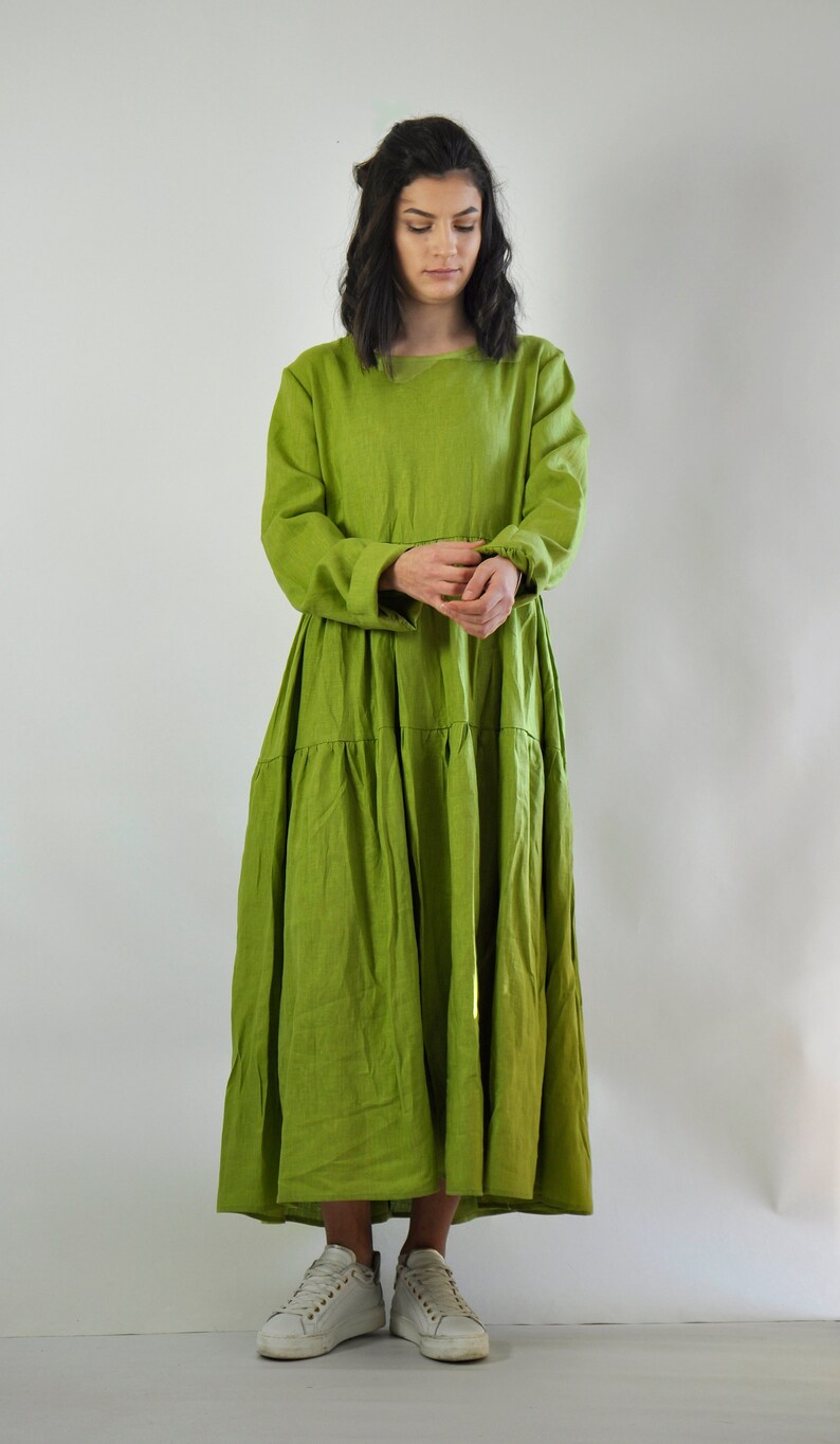 May include: A long, tiered, green linen dress with a round neckline and long sleeves. The dress is loose-fitting and has a flowy silhouette. The model is wearing white sneakers.