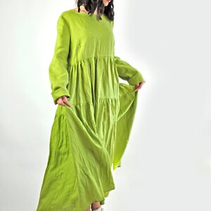 May include: A woman wearing a long, flowing green linen dress with a tiered skirt. She is wearing white sneakers and has her hair pulled back.