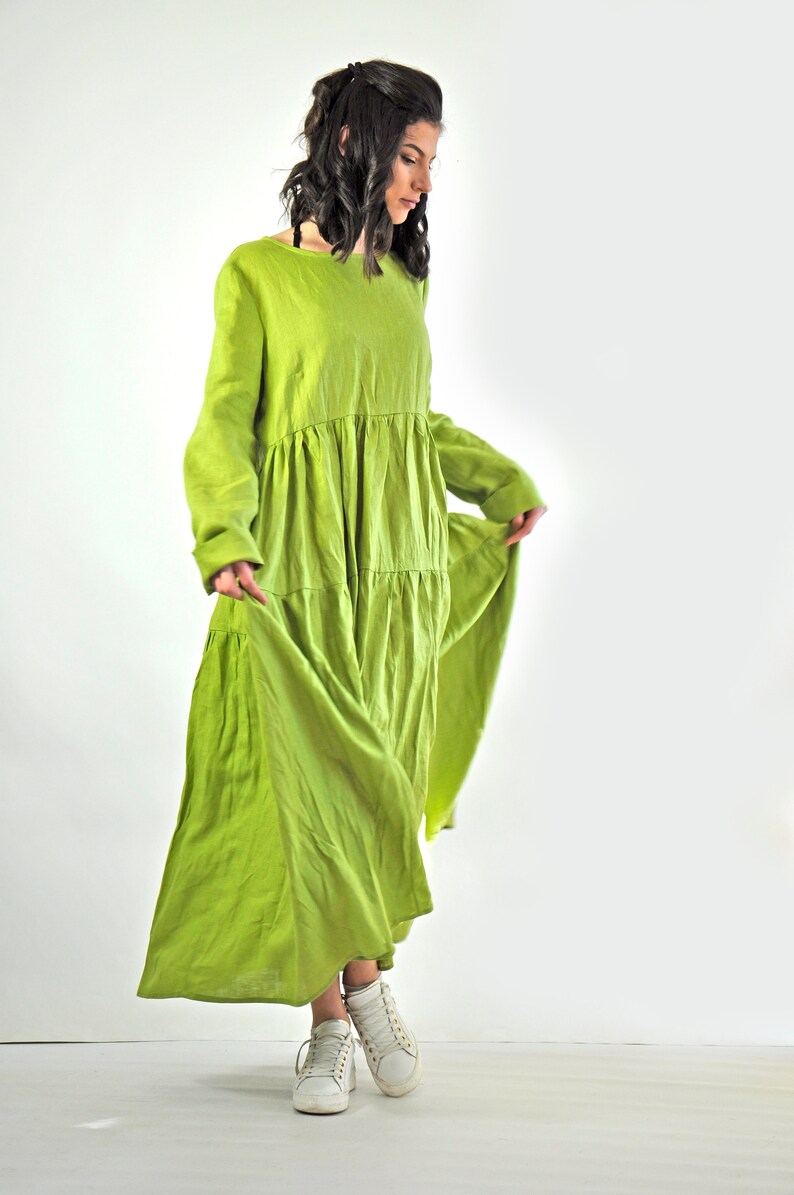 May include: A woman wearing a long, flowing green linen dress with a tiered skirt. She is wearing white sneakers and has her hair pulled back.