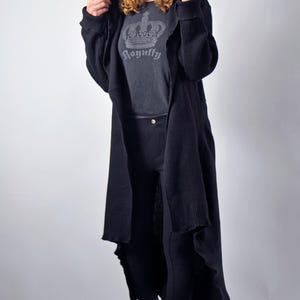 A model standing with the hood pulled up on a long black open-front cardigan, revealing a grey graphic t-shirt underneath.