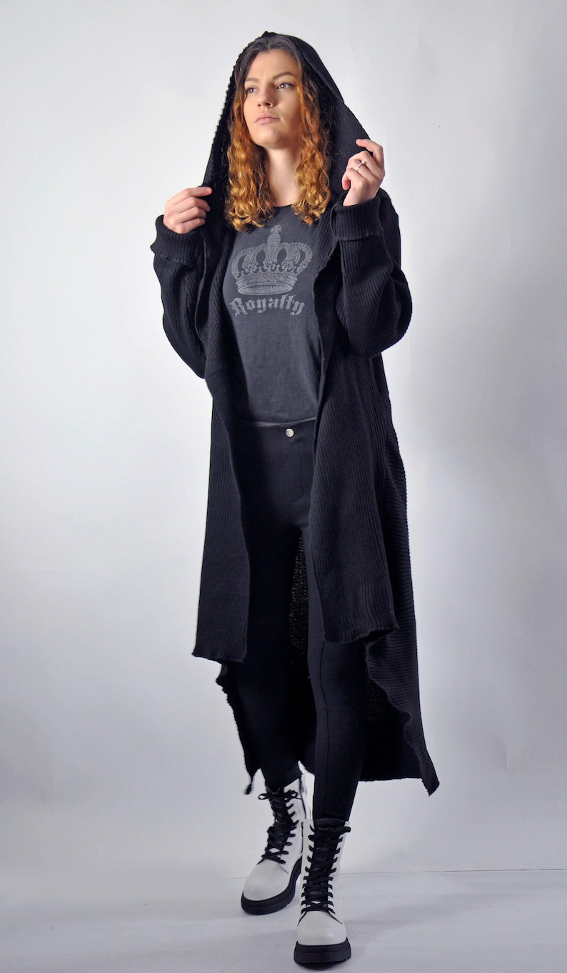 A model standing with the hood pulled up on a long black open-front cardigan, revealing a grey graphic t-shirt underneath.