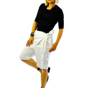 May include: A person wearing a black top, white wrap shorts, and black platform sandals. They are also wearing black sunglasses and have blonde hair. The outfit is set against a white background.