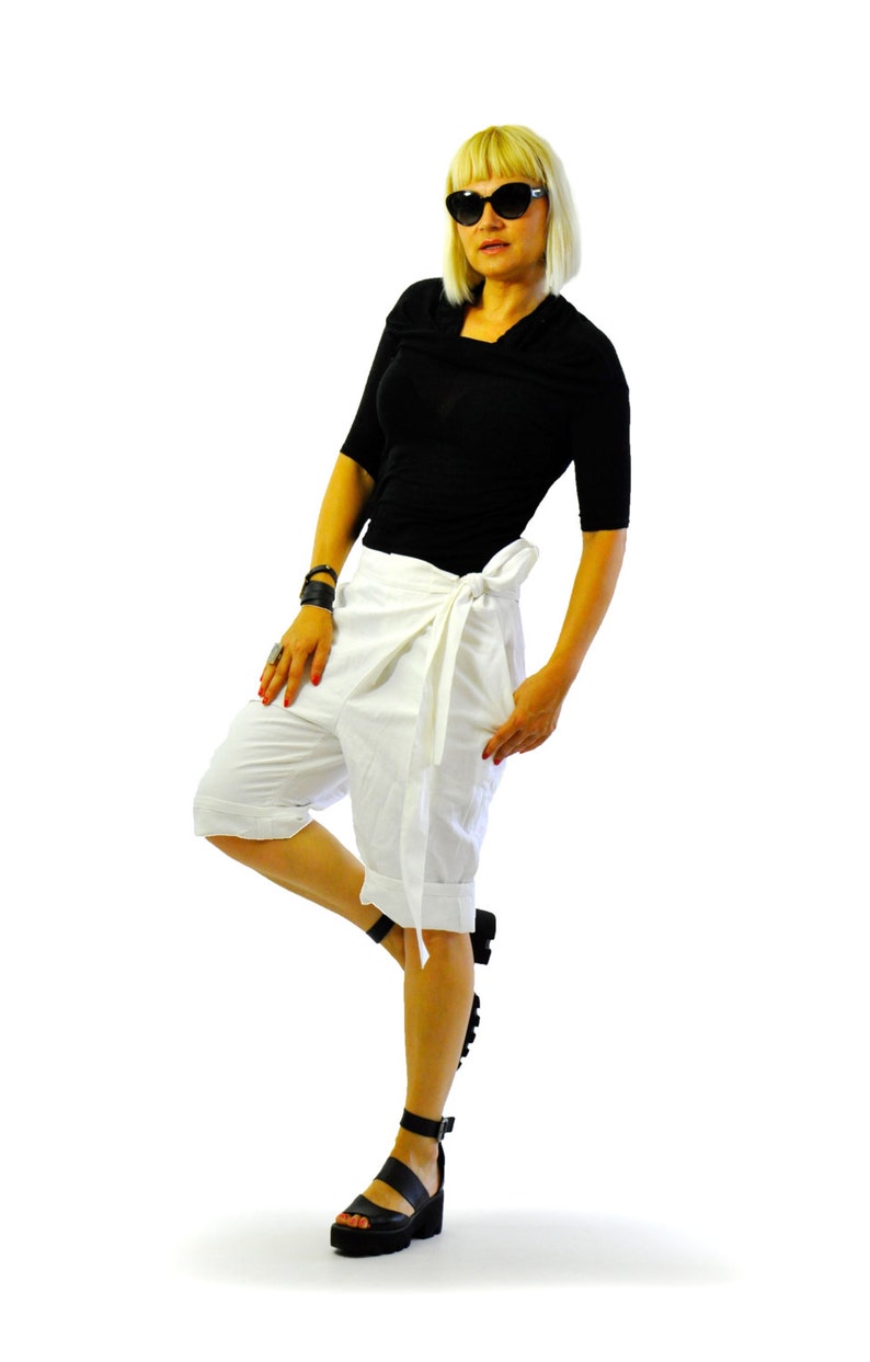 May include: A person wearing a black top, white wrap shorts, and black platform sandals. They are also wearing black sunglasses and have blonde hair. The outfit is set against a white background.