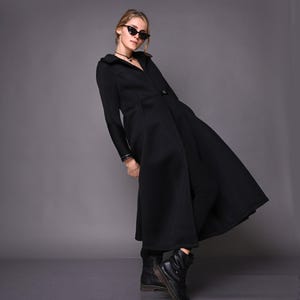 A model leaning to the side in a long black trench coat, showing the draped maxi skirt and black boots.