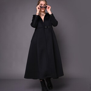May include: A woman wearing a black long-sleeved coat with a belt and black boots. She is holding a pair of sunglasses in front of her face.