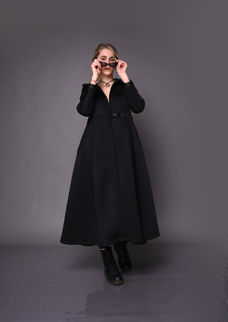 May include: A woman wearing a black long-sleeved coat with a belt and black boots. She is holding a pair of sunglasses in front of her face.