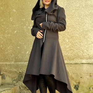 May include: A dark gray long-sleeved coat featuring an oversized hood, a high-low hemline, and a prominent white zipper closure running down the front. The coat has long sleeves with extended cuffs and a structured, flared silhouette that falls below the knees.