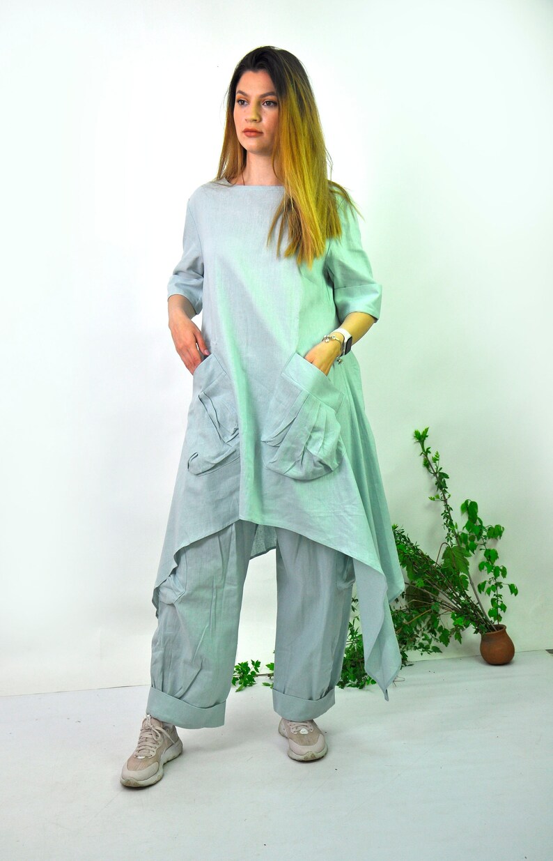 May include: A pale blue linen outfit, comprising a tunic top with an asymmetrical hem and large pockets, and matching wide-leg trousers. The model wears light-coloured trainers. The outfit is suitable for casual wear.