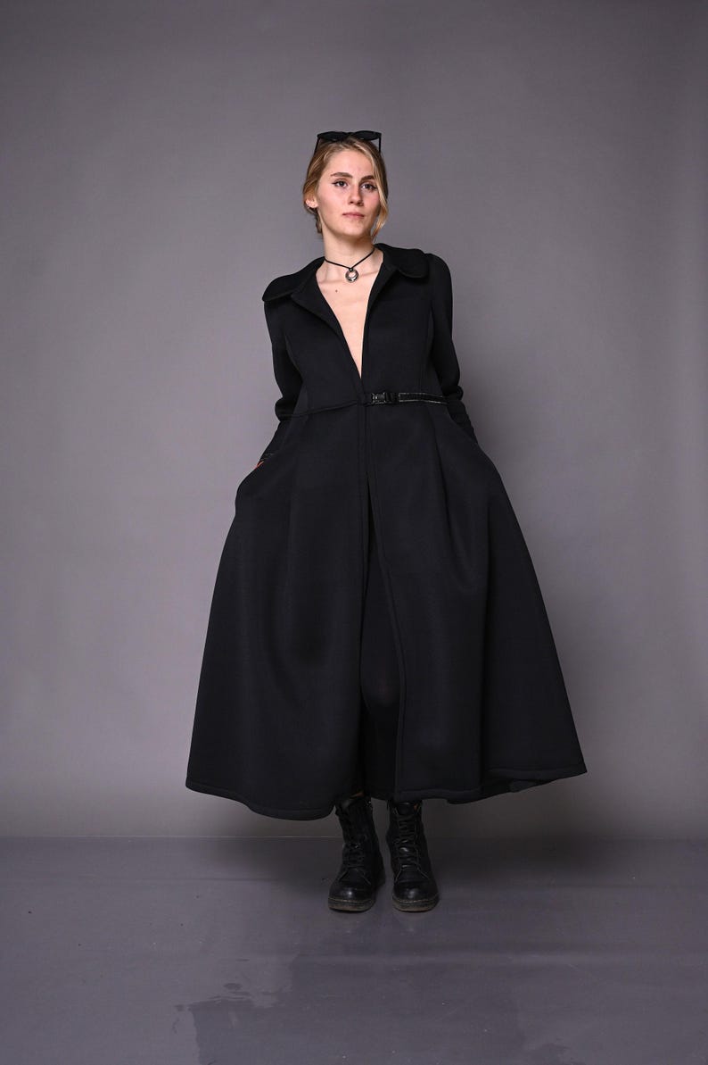 A model standing straight in a long black winter coat with a flared skirt and black boots against a grey background.