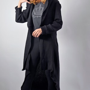 A full-length front view of a woman wearing a black maxi cardigan with a hood, layered over a grey shirt and black leggings.