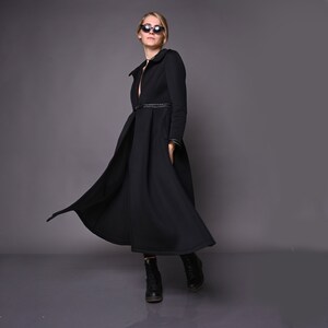 May include: A black long-sleeved dress with a fitted bodice and a flowing skirt. The dress has a belt at the waist and a small collar. The model is wearing black sunglasses and black boots.