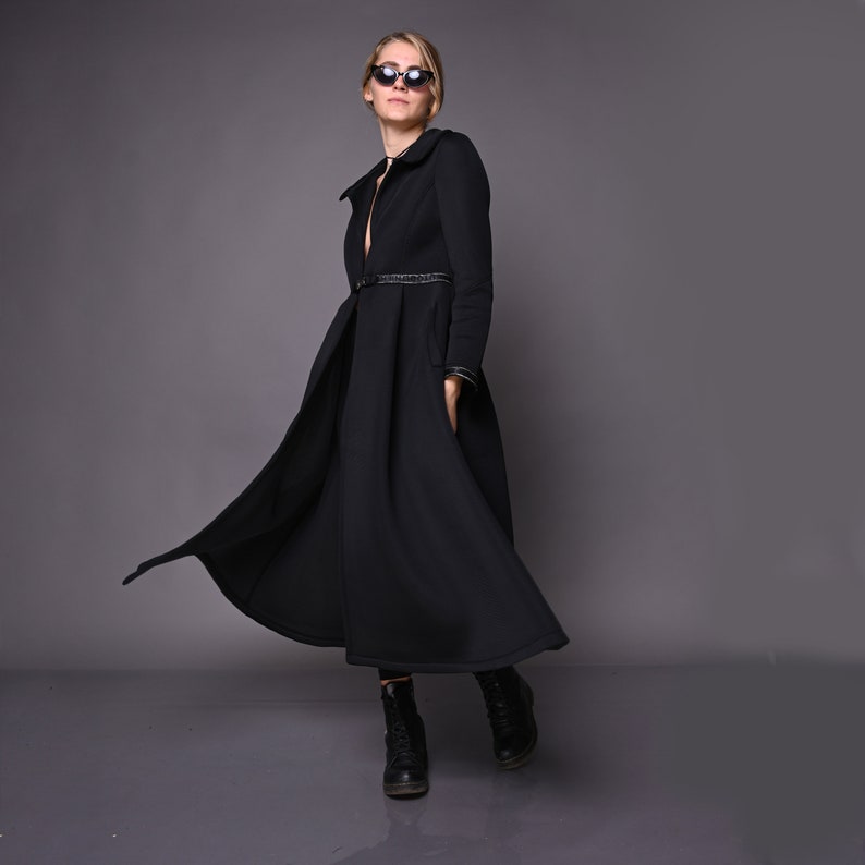 May include: A black long-sleeved dress with a fitted bodice and a flowing skirt. The dress has a belt at the waist and a small collar. The model is wearing black sunglasses and black boots.