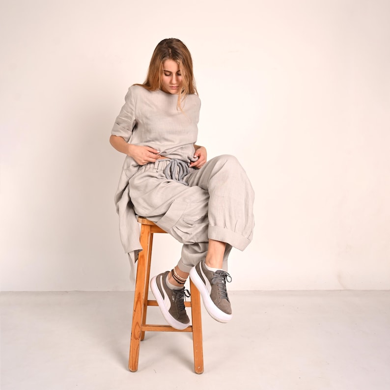 A woman with long blonde hair sits on a wooden stool, looking down at her light gray linen tunic and matching wide-leg pants with rolled cuffs, wearing gray sneakers.