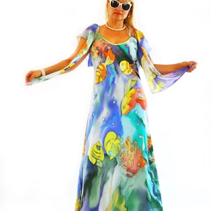 Puede incluir: Un vestido largo sin mangas con una vibrante escena submarina con patrones de peces coloridos, corales y algas en tonos azules, amarillos y naranjas. La prenda tiene un escote en V y mangas fluidas y transparentes. El texto gabyga es visible.