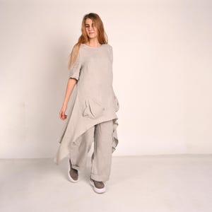 May include: A light grey linen outfit. The tunic top has an asymmetrical hem and pockets, and is paired with wide-leg trousers. The outfit is made of a soft, flowing fabric. The model is wearing grey trainers.