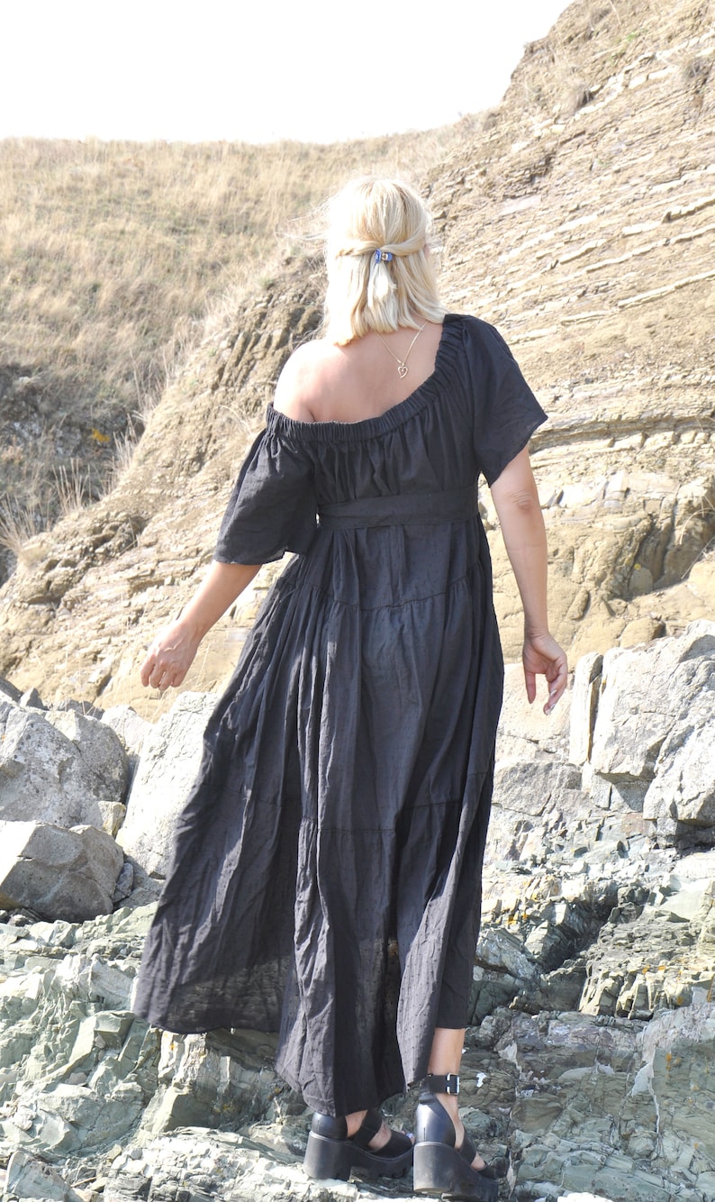 May include: A woman wearing a black off-the-shoulder maxi dress with a tiered skirt, standing on a rocky beach.