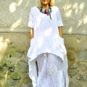 A woman in a white linen tunic and pants set, with a colorful fabric necklace, stands with hands in the tunic's pockets, looking at the camera.