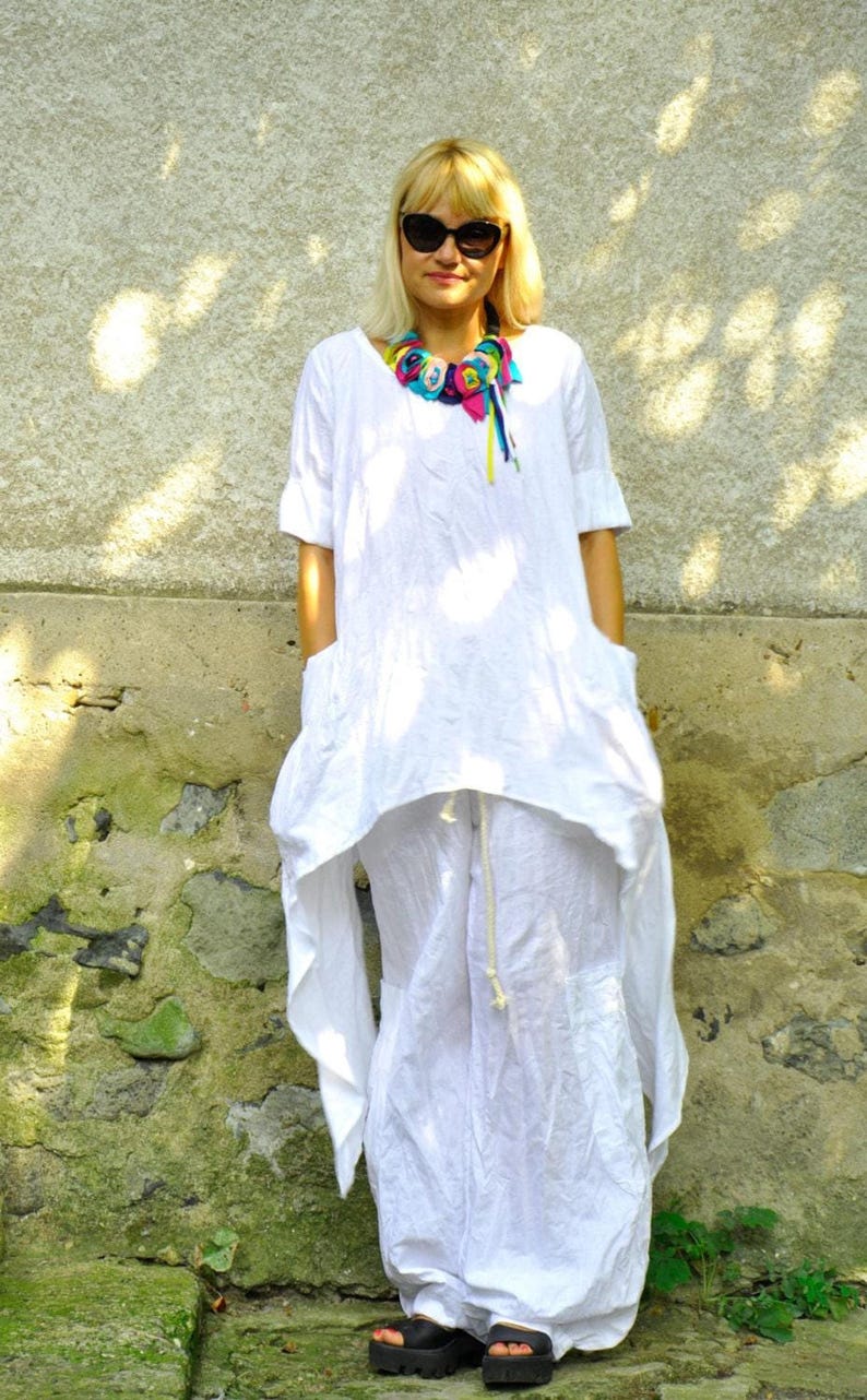 A woman in a white linen tunic and pants set, with a colorful fabric necklace, stands with hands in the tunic's pockets, looking at the camera.