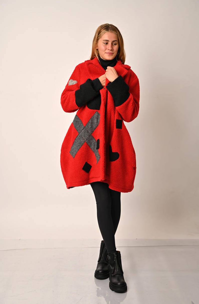 May include: Red fleece coat with black and gray geometric patches. The coat is knee-length and has a relaxed fit.  Features include a hood and long sleeves with black cuffs.  The patches are in the shapes of hearts and an 'X'.  The coat is shown on a mannequin against a white background.