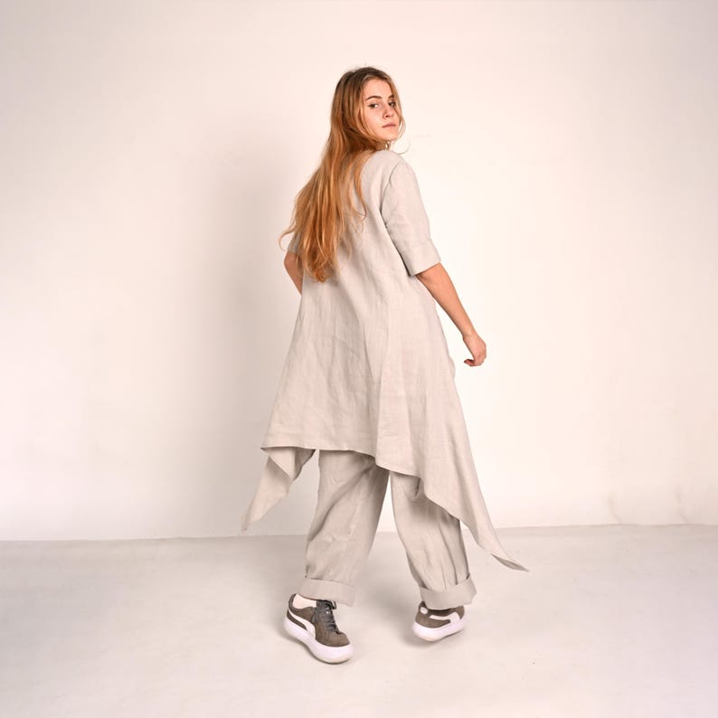 A woman with long blonde hair turns her head to look over her shoulder, wearing a light gray linen tunic with an asymmetrical hem and matching wide-leg pants, with gray sneakers.