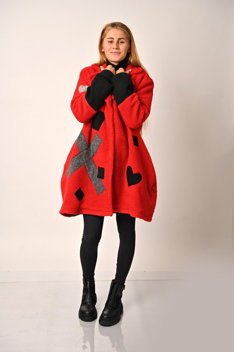 May include: A red, black and grey patterned fleece coat with a hood. The coat has a loose fit and is being worn by a person with long blonde hair. The person is wearing black leggings and black boots.