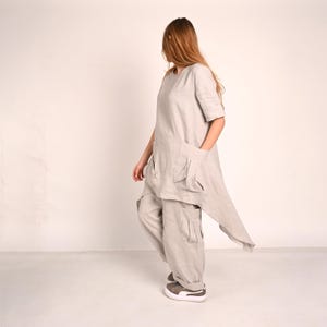 A woman with long blonde hair stands in profile, wearing a light gray linen tunic with large front pockets and an asymmetrical hem, paired with matching wide-leg pants with cargo pockets and gray sneakers.