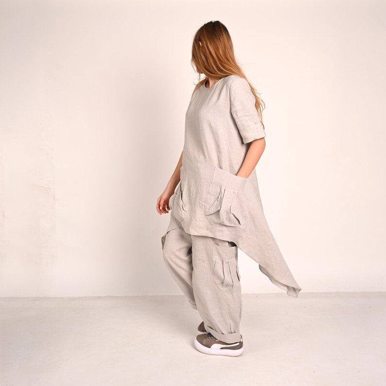 A woman with long blonde hair stands in profile, wearing a light gray linen tunic with large front pockets and an asymmetrical hem, paired with matching wide-leg pants with cargo pockets and gray sneakers.