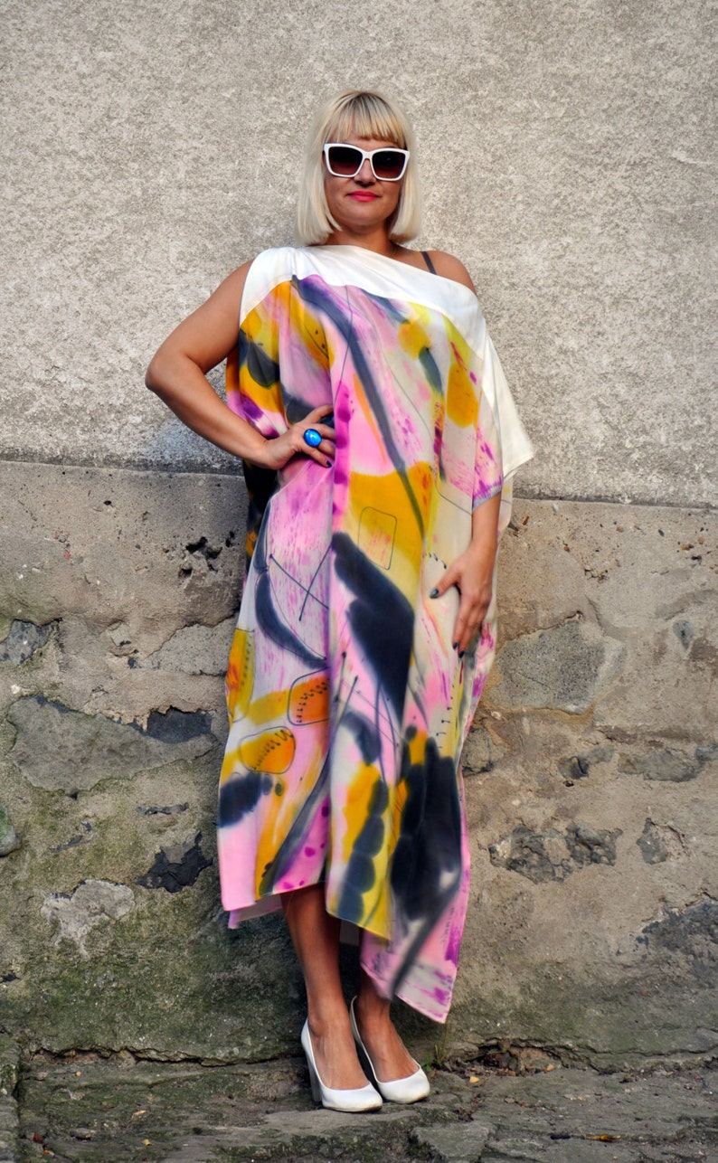 Puede incluir: Una mujer con un vestido de estampado abstracto blanco y rosa con un dise&ntilde;o de un solo hombro. Lleva gafas de sol blancas y un anillo azul.