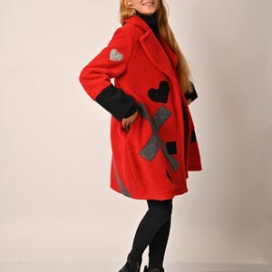 May include: A red fleece coat with black and grey patches in the shape of hearts and an X. The coat has a collar and long sleeves.