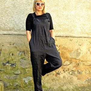 May include: A black jumpsuit with a bib-style top and wide legs. The jumpsuit has large pockets and button details on the straps. The person is wearing black sandals and white sunglasses. The background is a textured stone wall.