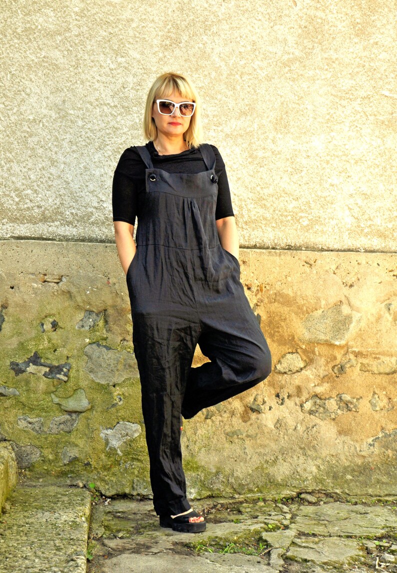 May include: A black jumpsuit with a bib-style top and wide legs. The jumpsuit has large pockets and button details on the straps. The person is wearing black sandals and white sunglasses. The background is a textured stone wall.
