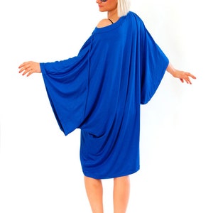 May include: A vibrant blue dress with wide sleeves and a draped design. The dress is paired with white sneakers. The model is wearing blue sunglasses. The dress has an off-the-shoulder neckline.