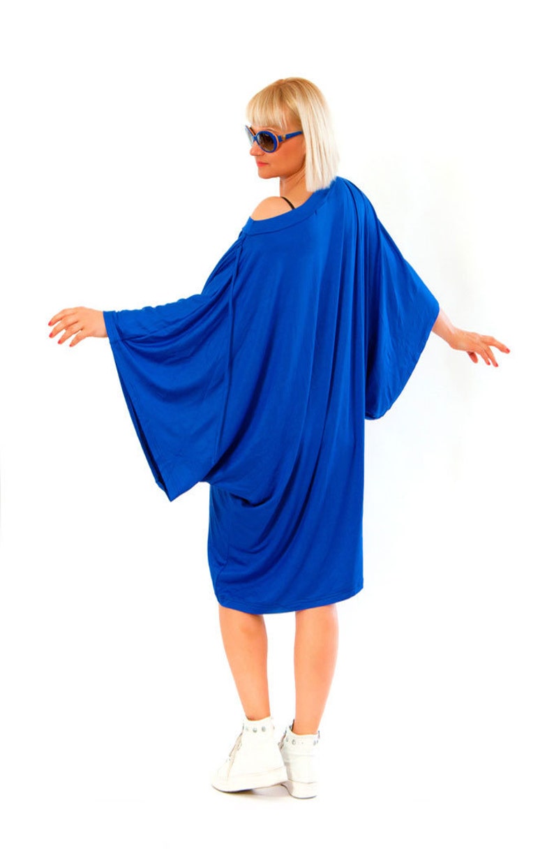 May include: A vibrant blue dress with wide sleeves and a draped design. The dress is paired with white sneakers. The model is wearing blue sunglasses. The dress has an off-the-shoulder neckline.