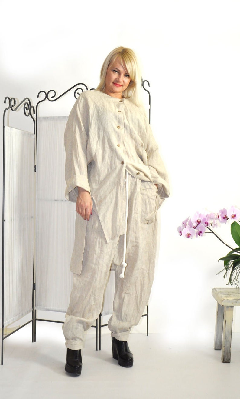 K&ouml;nnte beinhalten: Ein beiges Leinen-Outfit, bestehend aus einem Hemd mit Kn&ouml;pfen und einer locker sitzenden Hose, wird gezeigt. Das Outfit hat eine entspannte Passform und wird mit schwarzen Plateau-Stiefeln kombiniert. Ein wei&szlig;er Kordelzug ist an der Taille zu sehen. Der Hintergrund umfasst einen Raumteiler und einen kleinen Holzschemel.