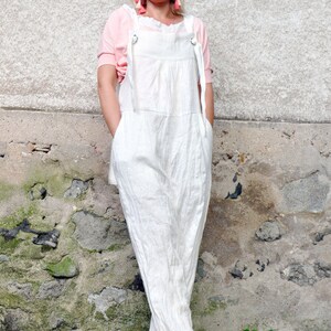 May include: A white linen jumpsuit with adjustable straps and a relaxed fit. The jumpsuit features a bib-style top and wide legs. The model is wearing a pink top underneath and sandals. The background is a textured stone wall.